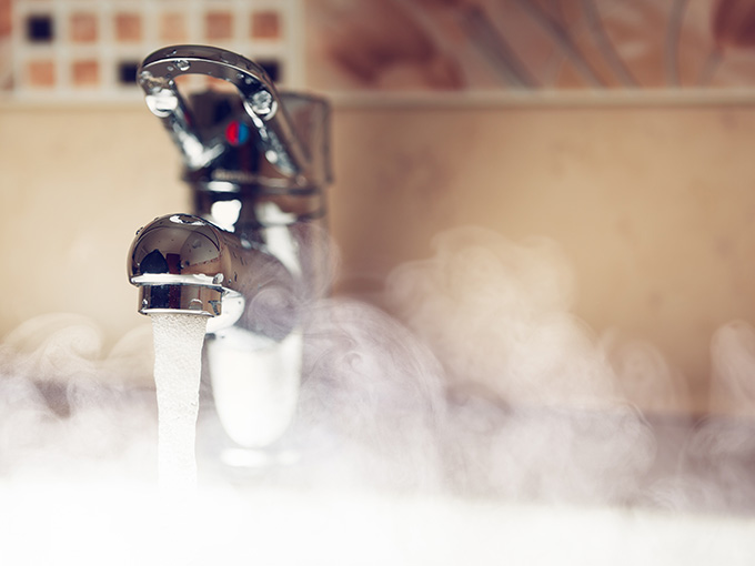 Nahaufnahme eines Wasserhahns in einer Badumgebung, aus dem warmes Wasser fließt und Wasserdampf aufsteigt